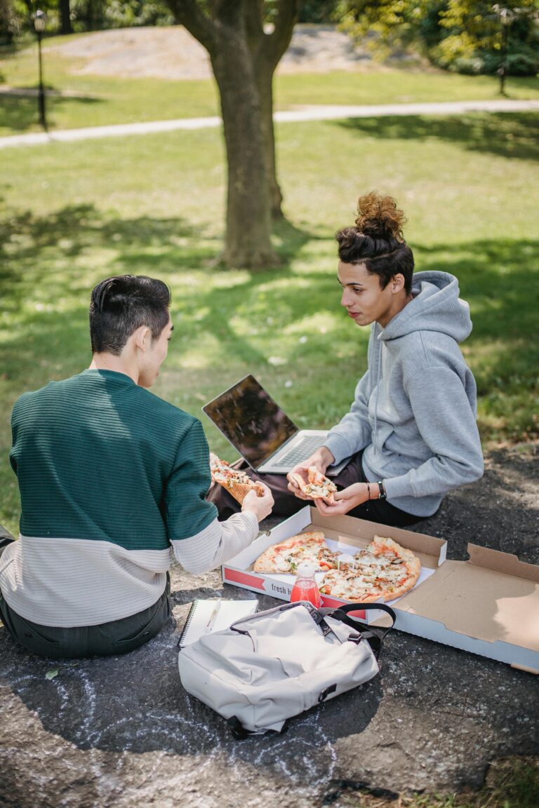 Two young men enjoying pizza and studying with a laptop in a park setting, embracing student life.