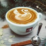 Cup of cappuccino with intricate swan latte art surrounded by seasonal spices and decorations.